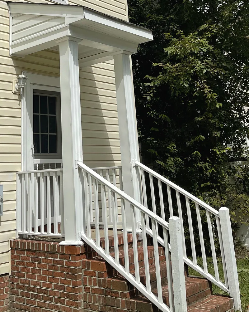 Side entry of a cream two-storey house, with a small portico and a white railing down brick steps